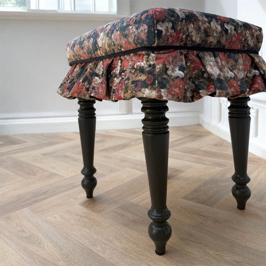 A vintage-style vanity stool with a floral upholstered cushion and ruffled skirt, supported by four turned black wooden legs, set on a light wood herringbone floor.