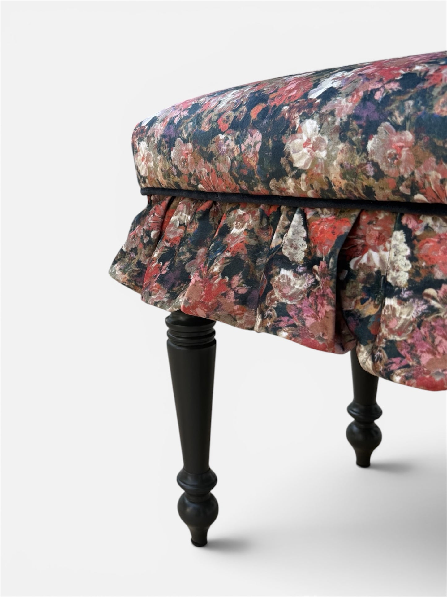 Close-up of an upholstered bench with a dark floral pattern cushion, pleated skirt, and turned black legs on a light gray background.
