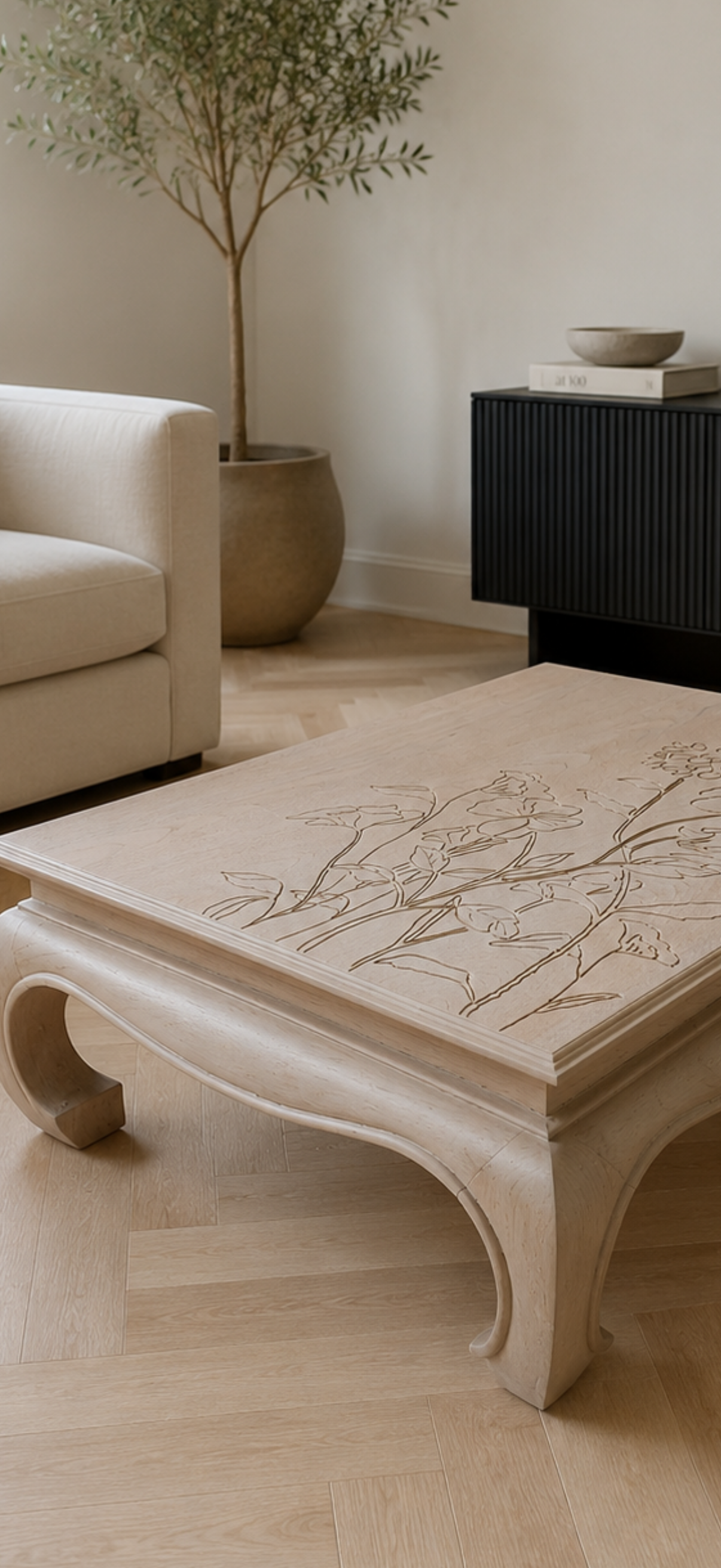 A light beige carved wooden coffee table with delicate floral engravings, set in a minimalist living room with a cream sofa and a black ribbed sideboard in the background.