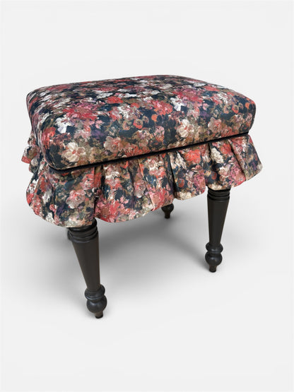A vintage-style upholstered stool with a ruffled skirt and painted turned legs, featuring a dense floral pattern in red, pink and cream on a black base.