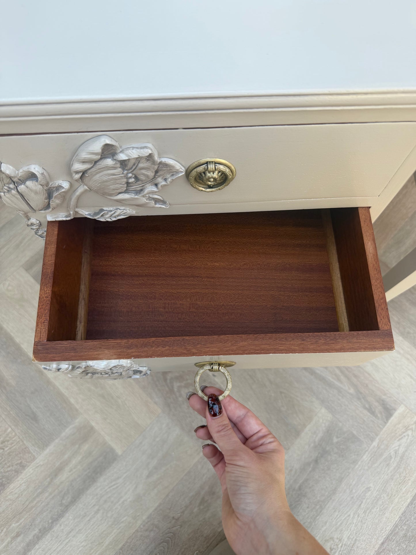 A close-up of a cream-colored wooden cabinet drawer with ornate gold floral handles and a brass ring pull, shown with a hand opening the drawer on a light wood herringbone floor.