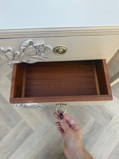 A close-up of a cream-colored wooden cabinet drawer with ornate gold floral handles and a brass ring pull, shown with a hand opening the drawer on a light wood herringbone floor.