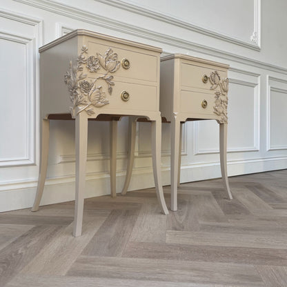 Two matching cream-colored nightstands with ornate floral carvings on the drawer fronts and gold-tone round knobs, set against a white paneled wall on a herringbone wood floor.