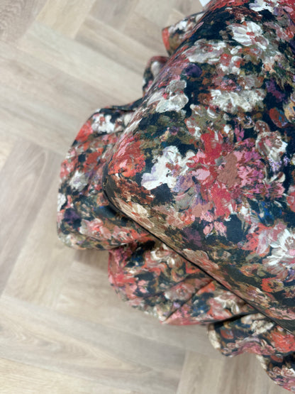 A close-up of a soft velvet, ruffled fabric with a dense floral pattern in red, white, black, and brown, laid out diagonally on a light wood parquet floor.
